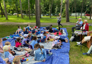Dzieci leżą w parku na niebieskiej macie słuchają babci czytającej książki